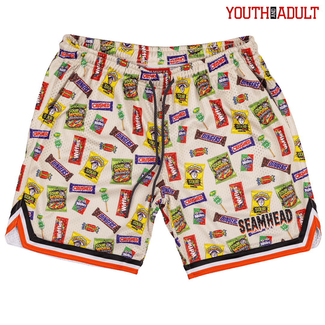 A pair of Halloween youth/adult lifestyle baseball 101 mesh shorts featuring an all-over print of various candy and snack packages on a cream mesh fabric, with orange, black, and white striped trim along the hem. The brand 'SEAMHEAD' is printed on the front.